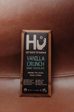 A chocolate bar wrapped in brown packaging with a focus on a square label featuring the brand name Hu and the message 'Get Back to Human.' The product is labeled as Vanilla Crunch Dark Chocolate with 70% organic cacao, vegan, primal, and free from refined sugars, dairy, soy, and other additives.