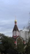 A traditional church with a distinct Russian architectural style, featuring a tall, pointed spire topped with a golden dome and cross. The building has a red and white color scheme and is partially obscured by trees. In the background, a modern, multi-story building is visible.