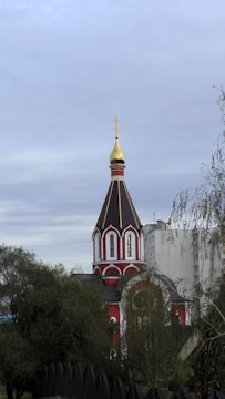 A traditional church with a distinct Russian architectural style, featuring a tall, pointed spire topped with a golden dome and cross. The building has a red and white color scheme and is partially obscured by trees. In the background, a modern, multi-story building is visible.