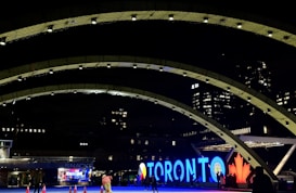 A night scene featuring illuminated, curved architectural structures in the foreground with a brightly lit sign spelling 'Toronto' in the background. The letters are large and prominently lit, accompanied by a red maple leaf. Several people are present, and there are traffic cones scattered on the ground. Modern buildings tower in the background, their windows lit up.