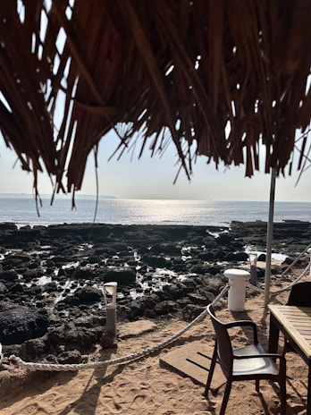 A coastal scene with a sandy foreground featuring a beach chair and a table. There is a rocky shoreline leading to a calm sea, with sunlight reflecting off the water. The top part of the image is framed by a thatched roof, adding a rustic element to the scene.