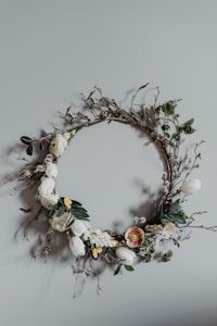 A decorative wreath made of various flowers and twigs, hanging on a plain light gray wall. The wreath features white and pale-colored flowers, green leaves, and delicate branches, arranged in a circular design.