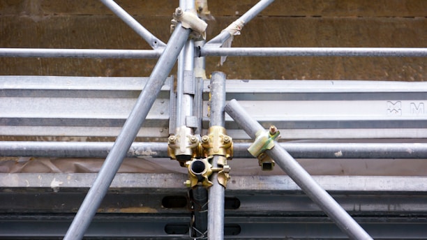A close-up view of a complex structure made of interlocking metal scaffolding tubes and clamps. The metal is predominantly silver and grey with hints of golden brass at the joints. The scaffolding appears sturdy and is likely used in construction or maintenance work.