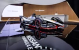 A sleek Formula E race car is prominently displayed in an elegant showroom with glossy black flooring that reflects the vehicle. The car features a sophisticated design with branding from companies such as Nissan, Richard Mille, and Michelin. A person stands behind a modern reception desk, enhancing the luxurious and technologically advanced atmosphere.