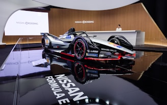 A sleek Formula E race car is prominently displayed in an elegant showroom with glossy black flooring that reflects the vehicle. The car features a sophisticated design with branding from companies such as Nissan, Richard Mille, and Michelin. A person stands behind a modern reception desk, enhancing the luxurious and technologically advanced atmosphere.