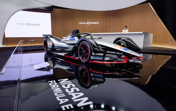 A sleek Formula E race car is prominently displayed in an elegant showroom with glossy black flooring that reflects the vehicle. The car features a sophisticated design with branding from companies such as Nissan, Richard Mille, and Michelin. A person stands behind a modern reception desk, enhancing the luxurious and technologically advanced atmosphere.