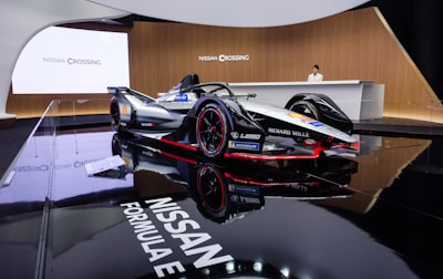 A sleek Formula E race car is prominently displayed in an elegant showroom with glossy black flooring that reflects the vehicle. The car features a sophisticated design with branding from companies such as Nissan, Richard Mille, and Michelin. A person stands behind a modern reception desk, enhancing the luxurious and technologically advanced atmosphere.