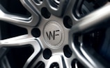 A close-up view of a polished metal car wheel hub with intricate design patterns. The central area features a circular logo with letters and text indicating manufacturing details. The surfaces are smooth and reflective, highlighting the craftsmanship of the metalwork.
