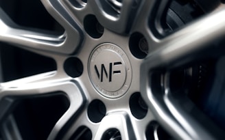 A close-up view of a polished metal car wheel hub with intricate design patterns. The central area features a circular logo with letters and text indicating manufacturing details. The surfaces are smooth and reflective, highlighting the craftsmanship of the metalwork.