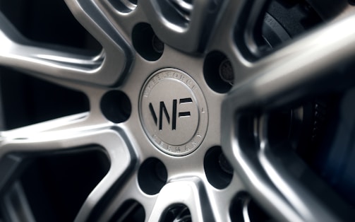 A close-up view of a polished metal car wheel hub with intricate design patterns. The central area features a circular logo with letters and text indicating manufacturing details. The surfaces are smooth and reflective, highlighting the craftsmanship of the metalwork.