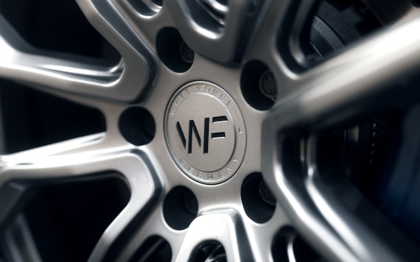 A close-up view of a polished metal car wheel hub with intricate design patterns. The central area features a circular logo with letters and text indicating manufacturing details. The surfaces are smooth and reflective, highlighting the craftsmanship of the metalwork.