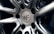 A close-up view of a polished metal car wheel hub with intricate design patterns. The central area features a circular logo with letters and text indicating manufacturing details. The surfaces are smooth and reflective, highlighting the craftsmanship of the metalwork.