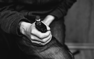 A person is holding a vape device in their hand, with visible details of a ring on their finger. The image is in black and white, and the person is wearing jeans and a dark sweater. The focus is on the hand and vaping device, with the background out of focus.