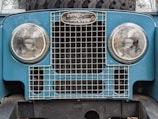 The front of a vintage Land Rover vehicle featuring two round headlights and a metal grille. The emblem above the grille displays the Land Rover logo. The vehicle's body is a light blue color and there is a spare tire mounted behind the grille.