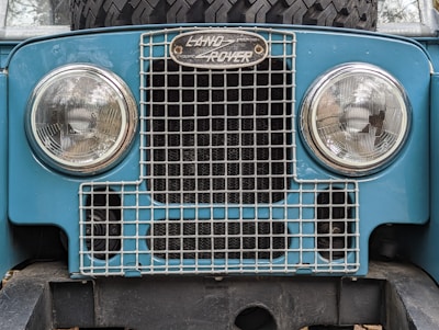 The front of a vintage Land Rover vehicle featuring two round headlights and a metal grille. The emblem above the grille displays the Land Rover logo. The vehicle's body is a light blue color and there is a spare tire mounted behind the grille.
