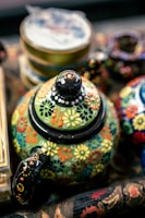 A colorful, intricately painted teapot is adorned with floral patterns in a variety of bright colors such as yellow, red, blue, and orange on a green background. The teapot sits among other blurred decorative items, suggesting a vibrant, artistic setting.