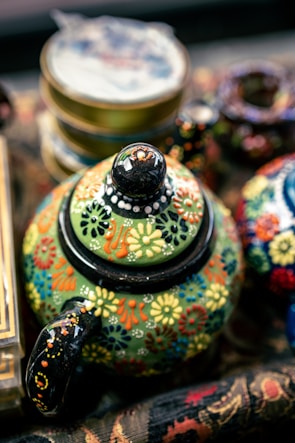 A colorful, intricately painted teapot is adorned with floral patterns in a variety of bright colors such as yellow, red, blue, and orange on a green background. The teapot sits among other blurred decorative items, suggesting a vibrant, artistic setting.