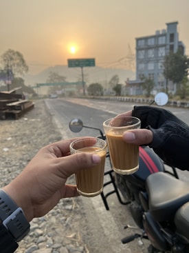 Two hands holding glasses of tea against the backdrop of an early morning setting with a rising sun. A motorcycle is parked nearby on the roadside, and a hazy sky suggests a cool or misty morning. Distant trees and a building are visible, with a quiet road stretching forward.