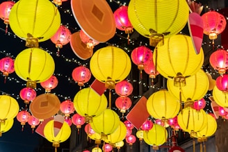 A collection of vivid, colorful paper lanterns hangs against a dark night sky. The lanterns are predominantly yellow and red with intricate designs and text on some of them. They are illuminated from within, casting a warm glow. Small twinkling lights are interspersed between the lanterns, enhancing the festive atmosphere.