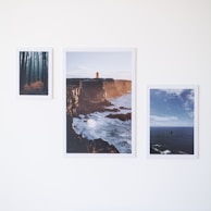 Three framed photographs are displayed on a white wall. The left photograph depicts a misty forest with tall trees and a path covered in fallen leaves. The center photograph showcases a rugged coastline with a large red lighthouse on a cliff, waves crashing against the rocks below. The right photograph features a vast ocean with a clear sky above and a small boat or rock formation in the distance.
