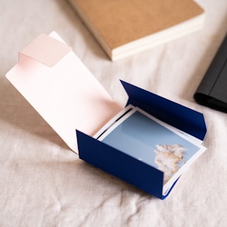 An open blue envelope or folder is laid out on a beige fabric surface. Inside, there are photographs, one showing a clear blue sky with some light-colored foliage or grass. Nearby are a couple of notebooks, one beige and the other black, suggesting a workspace or personal organization setting.