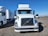 A smiling truck driver standing beside his parked truck in a spacious lot under clear blue skies.