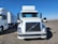 A smiling truck driver standing beside his parked truck in a spacious lot under clear blue skies.