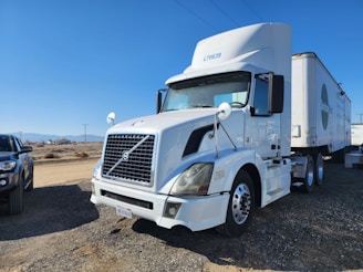 A large white semi-truck with a trailer is parked on a gravel surface, with clear blue sky and distant mountains in the background. Another vehicle, likely a pickup truck in a darker color, is partially visible on the left side.