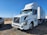 A large white semi-truck with a trailer is parked on a gravel surface, with clear blue sky and distant mountains in the background. Another vehicle, likely a pickup truck in a darker color, is partially visible on the left side.