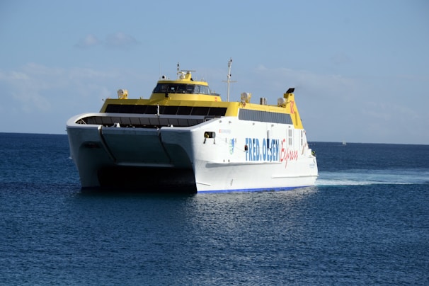 A large ferry with a white hull and yellow upper deck navigates through calm, blue ocean waters under a clear sky. The name 'FRED OLSEN Express' is visible on the side of the vessel.