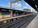 A railway station platform under a partly cloudy sky. The platform is empty, with tracks running parallel to it. The structure features a combination of brick and metal elements, with overhead electrical wires and a few utility poles visible in the background. The platform is well-lit with overhead lighting.