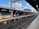 A railway station platform under a partly cloudy sky. The platform is empty, with tracks running parallel to it. The structure features a combination of brick and metal elements, with overhead electrical wires and a few utility poles visible in the background. The platform is well-lit with overhead lighting.