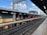 A railway station platform under a partly cloudy sky. The platform is empty, with tracks running parallel to it. The structure features a combination of brick and metal elements, with overhead electrical wires and a few utility poles visible in the background. The platform is well-lit with overhead lighting.