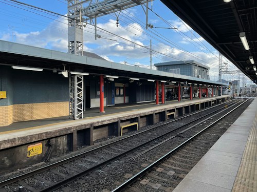 A railway station platform under a partly cloudy sky. The platform is empty, with tracks running parallel to it. The structure features a combination of brick and metal elements, with overhead electrical wires and a few utility poles visible in the background. The platform is well-lit with overhead lighting.