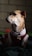 A small dog is wearing a red and white Christmas-themed outfit with a purple bell collar. The scene has low lighting, creating a shadowy background with hints of green and red objects.