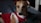 A dog wearing a red velvet outfit with white trim is resting on a dark-colored surface. The outfit has a festive appearance with a collar decorated with purple bells. The dog has a light brown coat and is looking towards the camera.