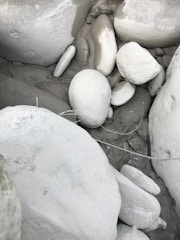 A collection of smooth, rounded white and grey stones clustered together. Some stones have subtle shades of beige and taupe in between, while a few strands of dried grass overlay the arrangement of rocks, suggesting a natural outdoor setting.
