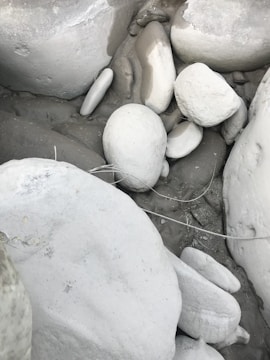 A collection of smooth, rounded white and grey stones clustered together. Some stones have subtle shades of beige and taupe in between, while a few strands of dried grass overlay the arrangement of rocks, suggesting a natural outdoor setting.