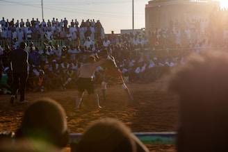 A vibrant wrestling match taking place outdoors, surrounded by a large crowd of spectators. The atmosphere is charged with excitement as people stand and sit around the dirt arena, intently watching the competitors. The sun is setting, casting a warm glow over the scene.