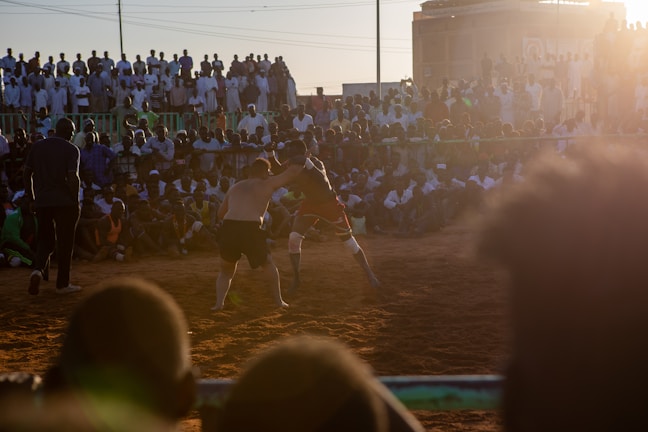 A vibrant wrestling match taking place outdoors, surrounded by a large crowd of spectators. The atmosphere is charged with excitement as people stand and sit around the dirt arena, intently watching the competitors. The sun is setting, casting a warm glow over the scene.