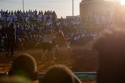 A vibrant wrestling match taking place outdoors, surrounded by a large crowd of spectators. The atmosphere is charged with excitement as people stand and sit around the dirt arena, intently watching the competitors. The sun is setting, casting a warm glow over the scene.