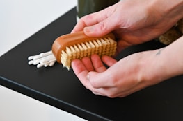 Hands are holding a wooden brush with natural bristles, positioned above a black surface. In the background, there are several cotton swabs partially visible.