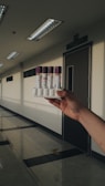 A hand holding a rack containing four vials with purple caps in a well-lit corridor. The corridor has a polished floor and several fluorescent light fixtures on the ceiling. A door with a sign above it is visible in the background.