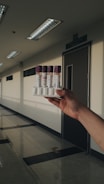 A hand holding a rack containing four vials with purple caps in a well-lit corridor. The corridor has a polished floor and several fluorescent light fixtures on the ceiling. A door with a sign above it is visible in the background.