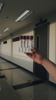 A hand holding a rack containing four vials with purple caps in a well-lit corridor. The corridor has a polished floor and several fluorescent light fixtures on the ceiling. A door with a sign above it is visible in the background.