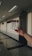 A hand holding a rack containing four vials with purple caps in a well-lit corridor. The corridor has a polished floor and several fluorescent light fixtures on the ceiling. A door with a sign above it is visible in the background.
