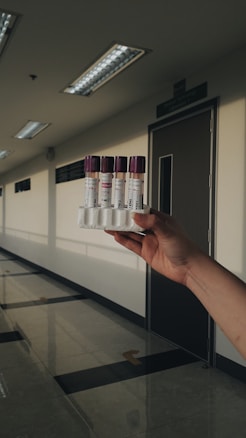 A hand holding a rack containing four vials with purple caps in a well-lit corridor. The corridor has a polished floor and several fluorescent light fixtures on the ceiling. A door with a sign above it is visible in the background.