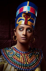 A person wears an elaborate headdress and colorful, beaded neckpiece, evoking ancient Egyptian style. The headdress is blue with golden and red designs, while the neckpiece features blue, red, and yellow beads. The individual has a serious expression and wears hoop earrings.