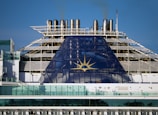 The image depicts the upper part of a cruise ship with smoke stacks and the ship's emblem prominently displayed. The structure is large and industrial, with a blue panel featuring a yellow sun-like symbol. Glass railings and other details of the ship's architecture are visible.