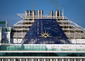 The image depicts the upper part of a cruise ship with smoke stacks and the ship's emblem prominently displayed. The structure is large and industrial, with a blue panel featuring a yellow sun-like symbol. Glass railings and other details of the ship's architecture are visible.